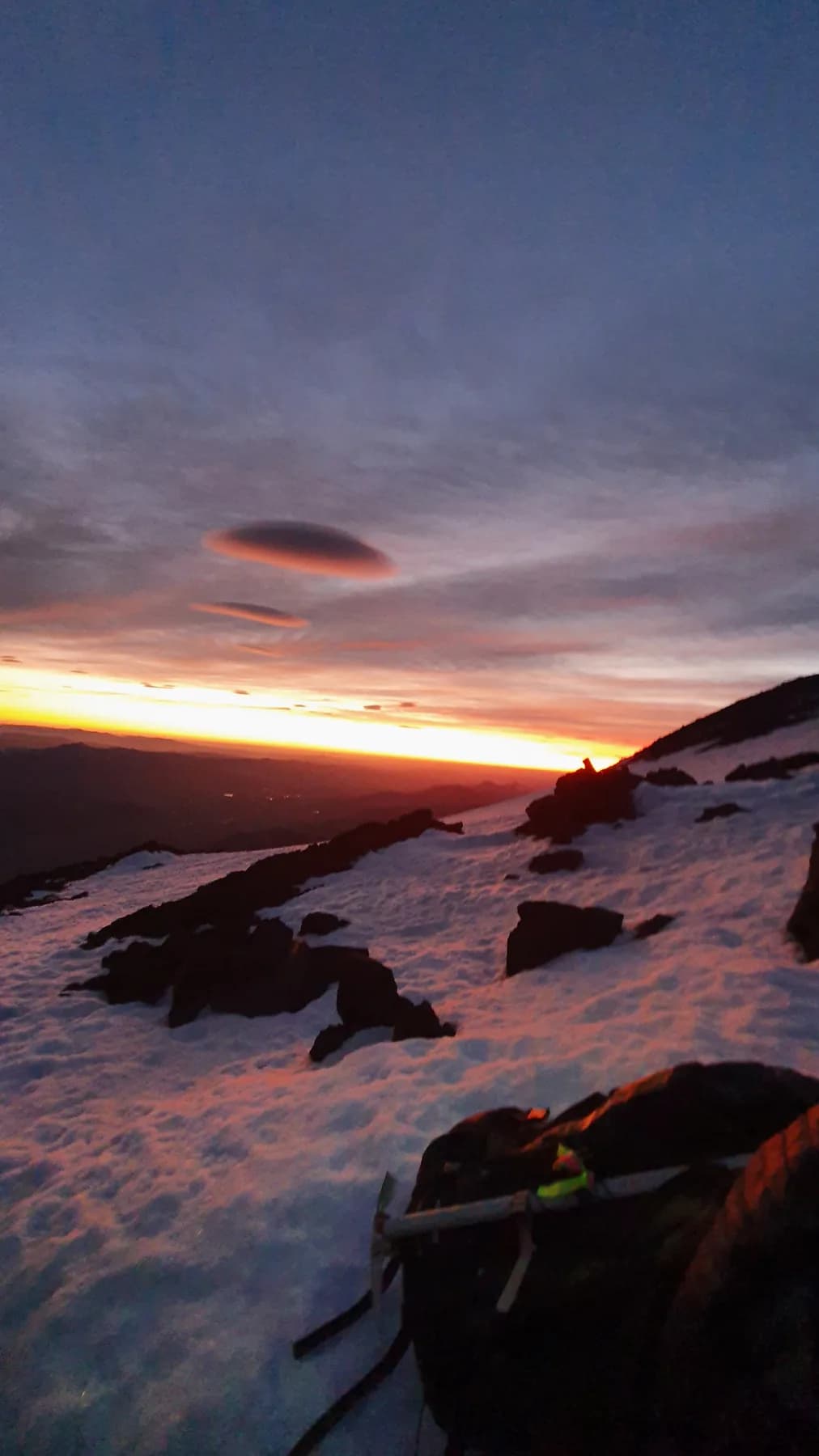 Sunrise from the upper slopes of Lanín, two lenticular clouds over the range, snow and volcanic rock in the foreground with an ice axe and pack