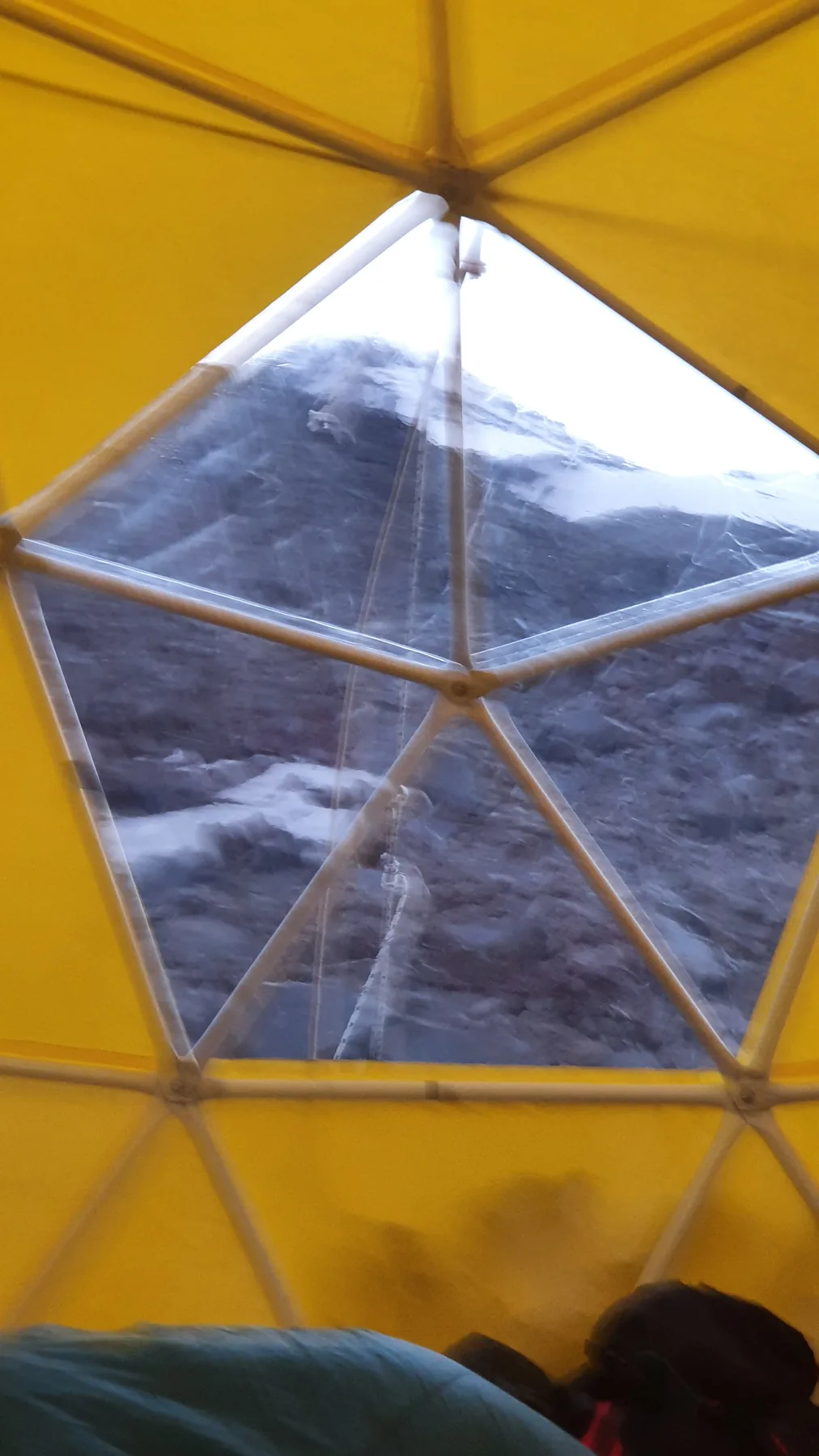 Looking up through the clear roof panel of the yellow geodesic refugio on Lanín at the snow slope and rock bands of the route above